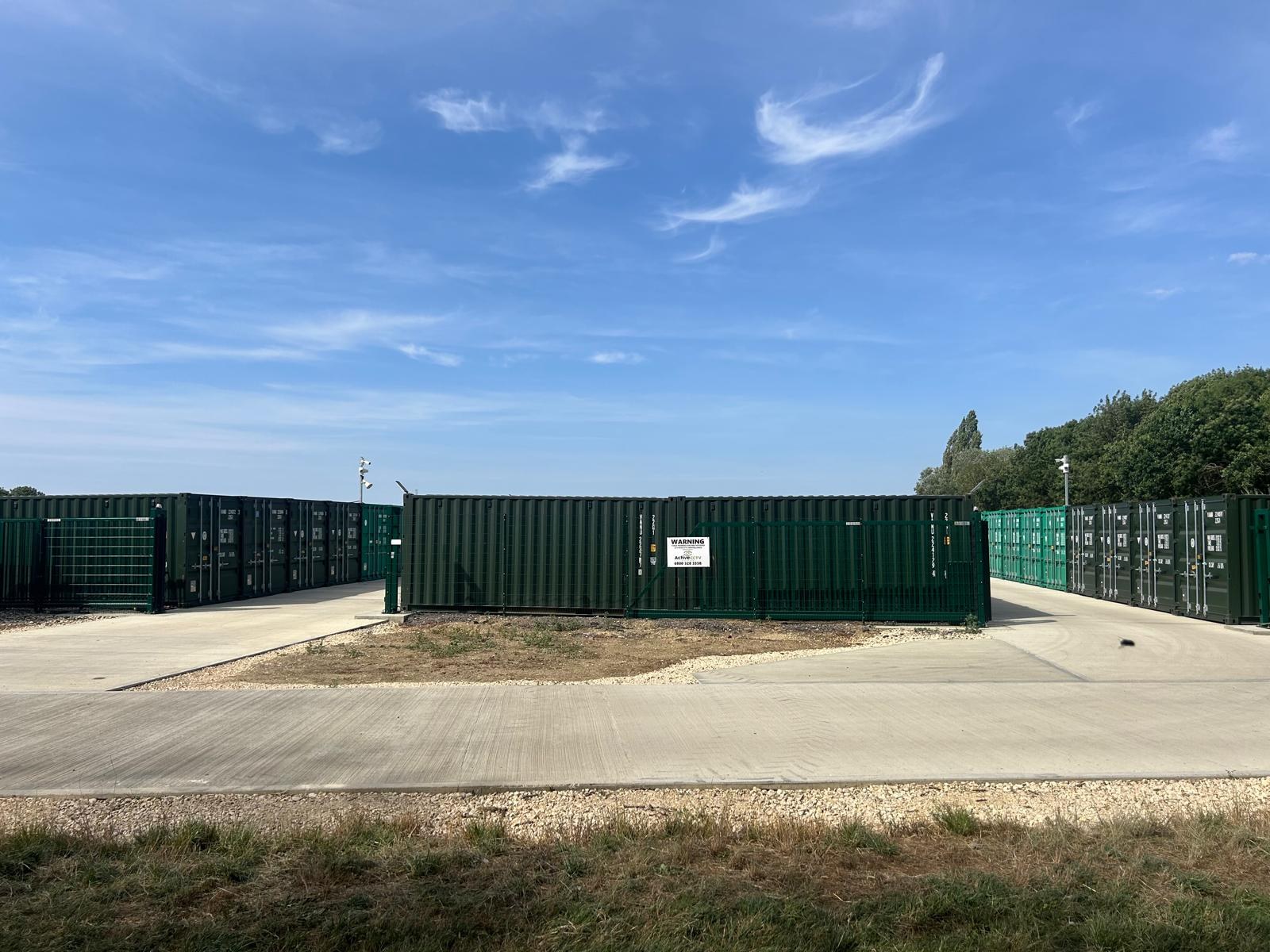 Rows of green container storage units behind mesh security gates, with an active CCTV warning sign
