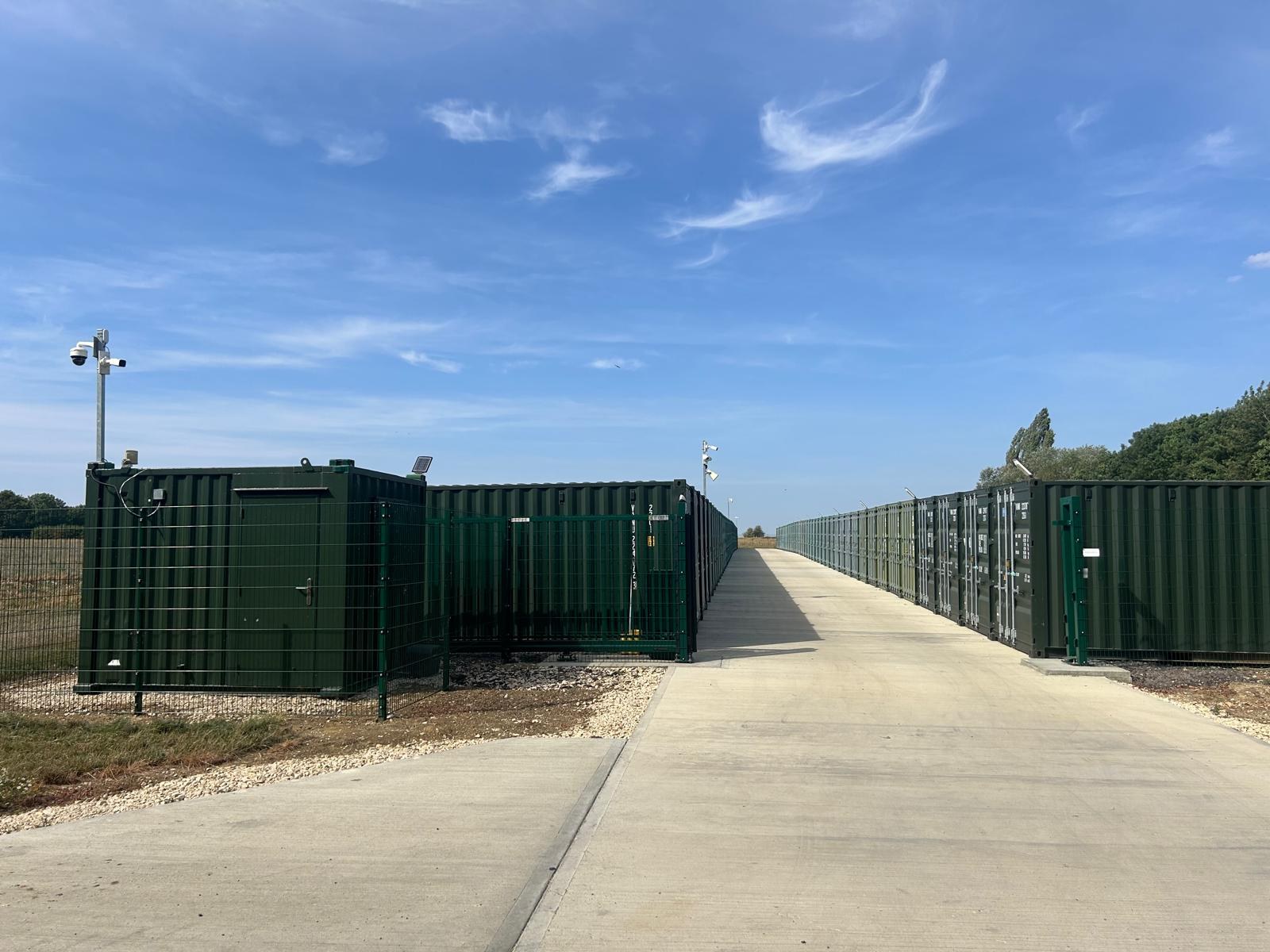 Green site container and CCTV mast beside long rows of container storage units on a concrete roadway