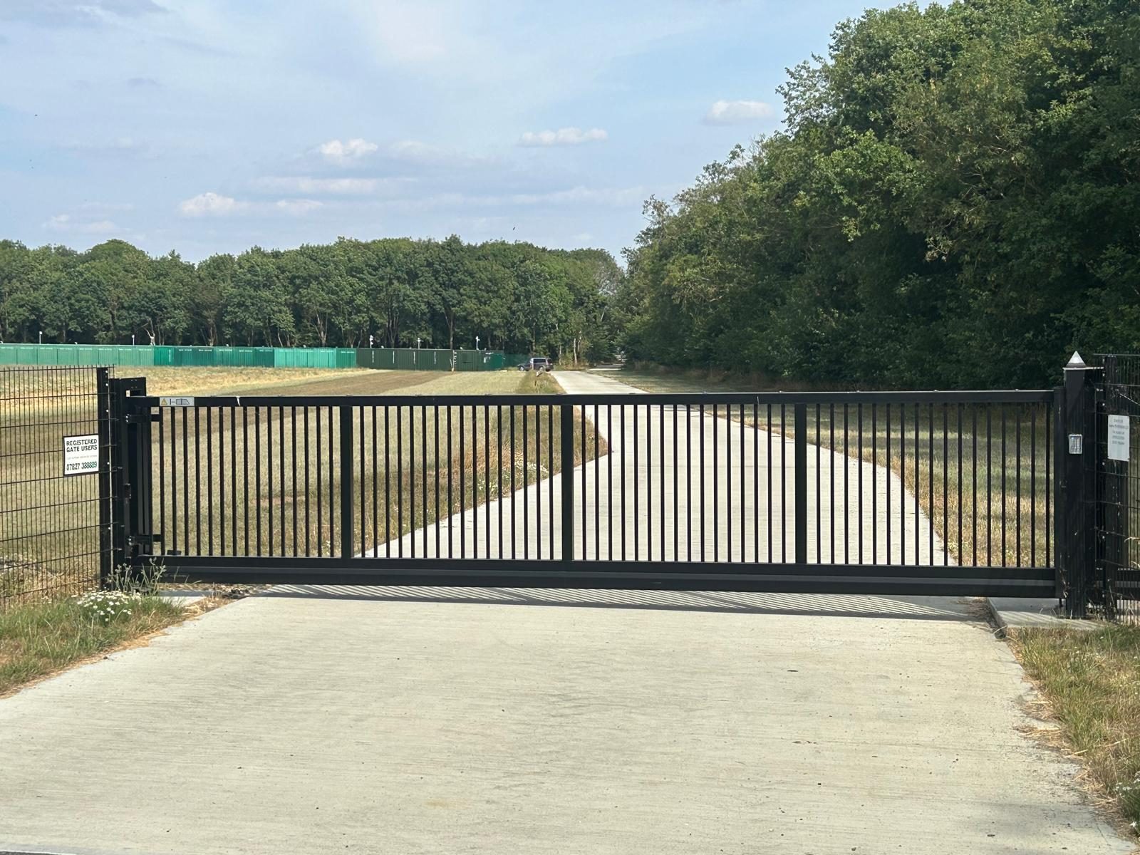 Black automated sliding gate closed across the concrete access road, with green storage containers in the distance